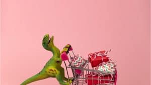 A toy dinosaur pushing a toy shopping cart filled with presents, against a pink background