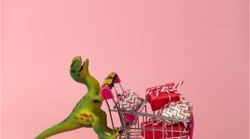 A toy dinosaur pushing a toy shopping cart filled with presents, against a pink background
