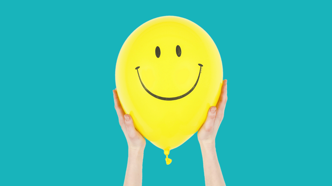 A yellow balloon is held between two hands. It has a smiley face drawn on it.